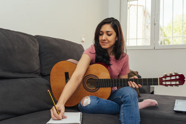 Mulher sentada no sofá segura violão e anota notas musicais