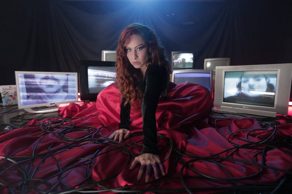 Pitty de vestido vermelho e sentada com TVs ao redor