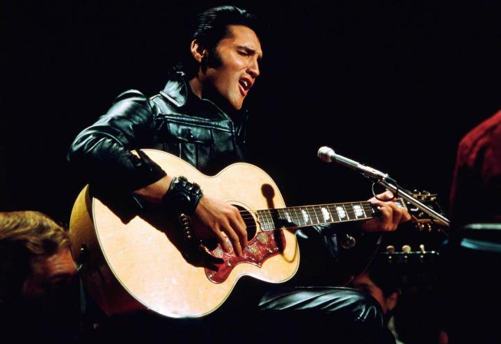 Elvis Presley, com seu traje preto, tocando violão e cantando durante especial de TV