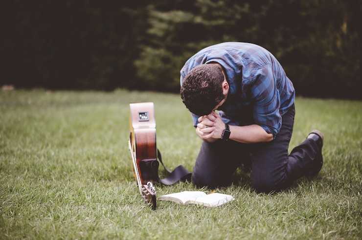 Homem ajoelhado em gramado rezando ao lado de violão