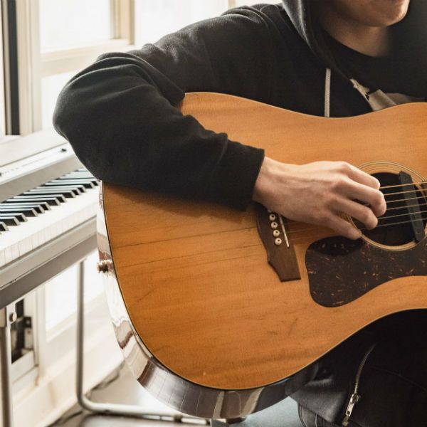 jovem-tocando-violao Jovem sentado no banco de um home studio toca violão. Atrás dele, há um teclado